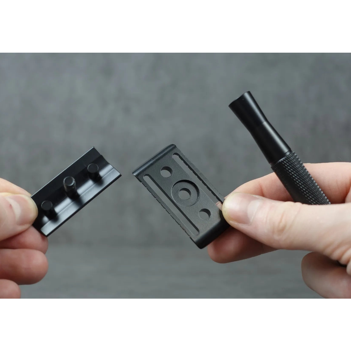 Two hands holding a black razor blade, razor head, and handle against a gray background